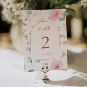Puede incluir: Una tarjeta con número de mesa con un diseño floral, que muestra la palabra "table" y el número "2" en rosa. La tarjeta presenta un patrón de acuarela de flores rosas, azules y amarillas. Los nombres "Cecilia & Harrison" y la fecha están impresos debajo.