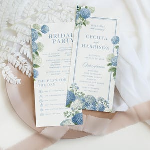 May include: Two wedding invitation cards with blue hydrangea floral designs. One card reads "Bridal Party" and the other reads "Cecilia and Harrison" with the order of service. The cards are on a pink plate with a white background.