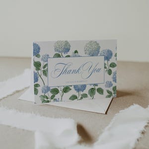 Peut inclure: Carte de remerciement pliée avec un motif floral d'hortensias. La carte présente une bordure bleue et les mots "Thank You" en écriture élégante. La carte est entourée de fleurs d'hortensias bleues et de feuilles vertes. Les noms "Cecilia & Harrison" sont imprimés.