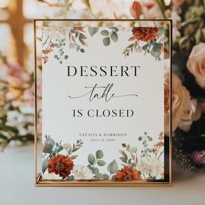 Puede incluir: Un letrero rectangular en un marco dorado con las palabras "DESSERT table IS CLOSED" en negro. El letrero está decorado con arreglos florales en tonos blancos, naranjas y verdes. Los nombres "CECILIA & HARRISON" y una fecha están en la parte inferior.