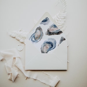 May include: A white envelope with a watercolor design of four oysters in shades of blue and gray. The envelope is open, revealing the oyster artwork. White ribbon and a wax seal with a floral design are also visible.