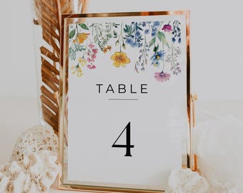 May include: A gold-framed table number card displaying the word "TABLE" and the number "4" in black. The card's top edge features a watercolour floral design with yellow, blue, and purple flowers. The card is on a white surface.