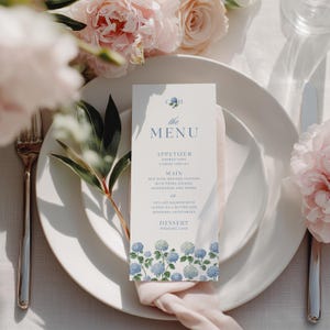May include: A wedding menu card with a floral design featuring blue hydrangeas at the bottom. The menu card is placed on a white plate with a pink napkin and a fork and knife. The menu includes the text "the MENU" and the menu items.