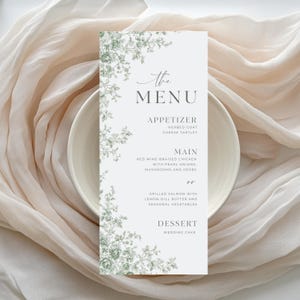 Pode incluir: Um cartão de menu de casamento com um design floral em tons de verde e branco. O menu inclui entradas, pratos principais e sobremesas. O cartão está colocado num prato branco e num tecido macio de cor creme.