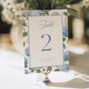 May include: A wedding table number card with a floral hydrangea design. The card features the word "Table" in script, the number "2", and the names "Cecilia & Harrison" with a date. The card is held in a silver metal holder.