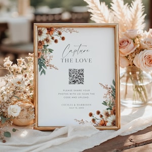 May include: A framed wedding sign with the text "Capture the Love" and a QR code. The sign is decorated with floral accents in shades of white, orange, and green. The sign also includes the names Cecilia & Harrison and the date July 15, 20XX.