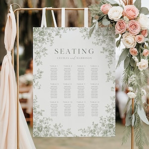 May include: A wedding seating plan featuring the names Cecilia and Harrison. The sign is white with a green floral border. Guest names are listed under table numbers. The sign is suspended from a gold metal stand, with floral decorations.
