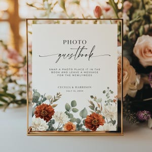 May include: A framed wedding sign with the words "Photo Guestbook" in elegant script. The sign includes instructions for guests and the names Cecilia & Harrison, with the date July 15, 20XX. Floral accents in white, rust, and green adorn the bottom.