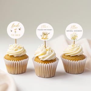 Puede incluir: Tres cupcakes con glaseado blanco y chispas de perlas coronados con adornos redondos de papel para cupcakes. Los adornos presentan un diseño de abejas y flores con el texto "Bride to Bee", "Ella's Found Her Honey" y "Ella's Bridal Shower".