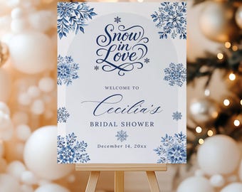 May include: A white sign on a wooden easel with the text "Snow in Love" in navy blue script, surrounded by blue floral snowflake designs. The sign welcomes guests to Cecilia's bridal shower, with the date December 14, 20xx.