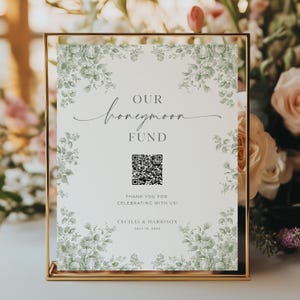 May include: A gold-framed sign with the words "OUR honeymoon FUND" in elegant script. The sign features a QR code and is decorated with a floral border. The text "THANK YOU FOR CELEBRATING WITH US!" and names are also visible.