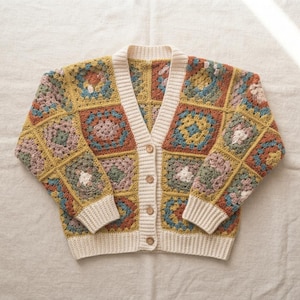 May include: A long-sleeve cardigan made of crocheted granny squares in shades of mustard yellow, terracotta, sage green, and dusty pink. The garment features a cream-coloured ribbed V-neck collar, cuffs, and hem, secured by four small wooden buttons.