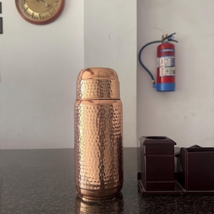 Puede incluir: Una botella de agua de color cobre con un diseño texturizado y martillado. La botella tiene una tapa a juego y se encuentra sobre una superficie oscura y moteada. Un reloj redondo y un extintor son visibles en el fondo.