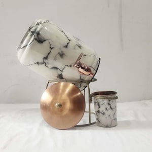 May include: A marble-patterned beverage dispenser with a copper spigot and lid, set on a metal stand. A matching marble cup is also present. The dispenser is designed for serving drinks.
