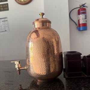 May include: A copper water dispenser with a hammered texture, a tap, and a lid with a small knob. The dispenser is on a dark countertop. A clock and a fire extinguisher are in the background.