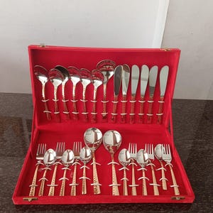 Puede incluir: Un juego completo de cubiertos, que incluye cucharas, tenedores y cuchillos, está cuidadosamente dispuesto dentro de un estuche de terciopelo rojo. La cubertería tiene un acabado plateado brillante con mangos decorativos dorados. El juego está diseñado para cenas formales.