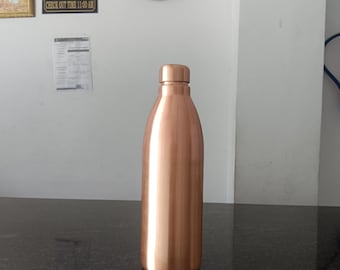 May include: A copper-colored water bottle with a rounded body and a screw-on cap. The bottle is standing on a dark, speckled surface. The background includes a clock and signs that say "RECEPTION", "CHECK IN TIME 12:00 PM", "CHECK OUT TIME 11:00 AM", "YOU ARE UNDER CAMERA SUPERVISION", and "NO SMOKING".