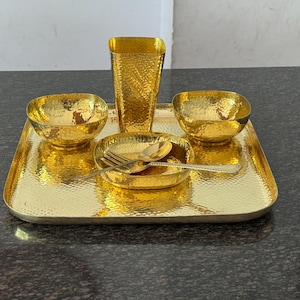 Puede incluir: Un juego de servir dorado martillado. El conjunto incluye una bandeja rectangular, dos cuencos, un plato pequeño con cuchara y tenedor, y un jarrón alto y cuadrado. Los artículos tienen una superficie texturizada y reflejan la luz.