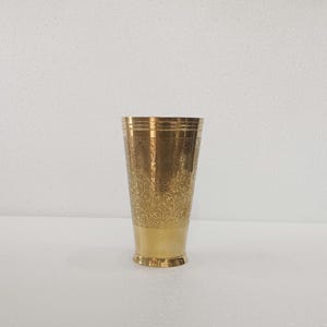 May include: A brass-colored drinking glass with an ornate floral pattern etched into the upper portion. The base of the glass is a solid brass color. The glass has a slightly flared shape and a decorative rim.