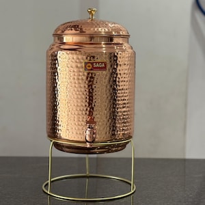 Op de afbeelding: Een koperen waterdispenser met een gehamerde textuur, een kraan en een deksel met een kleine knop. De dispenser staat op een goudkleurige metalen standaard. Het logo "SAGA" is zichtbaar aan de voorkant.
