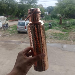Puede incluir: Una botella de agua de cobre con una textura martillada. La botella tiene una tapa de rosca con asa y cuello estrecho. El cobre tiene una superficie brillante y reflectante. La botella es sostenida por una persona.