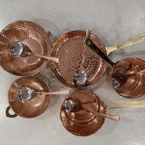 May include: Copper cookware set with hammered texture. Includes small pots with handles, a frying pan, and serving spoons. The copper colour contrasts with the silver spoons and brass handles.