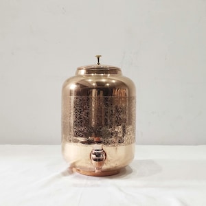Puede incluir: Un dispensador de agua de cobre con grifo y tapa. El dispensador tiene un patrón floral grabado alrededor del centro y un acabado pulido. El dispensador se asienta sobre una base de madera.