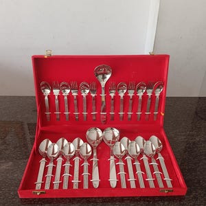 May include: A complete silver-colored flatware set displayed in a red velvet-lined case. The set includes spoons, forks, and a ladle, all with decorative handles. The case is open, showcasing the arrangement of the utensils.