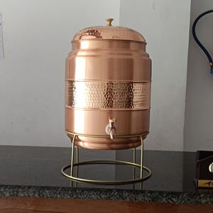 May include: A copper water dispenser with a hammered texture band. The dispenser has a tap and sits on a gold-coloured metal stand. The lid is also copper and has a small knob on top. The dispenser is cylindrical and has a polished finish.