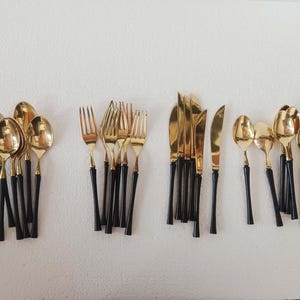 Puede incluir: Un conjunto de cubiertos dorados con mangos negros. El juego incluye cucharas, tenedores y cuchillos, dispuestos sobre una superficie blanca. Los utensilios tienen un acabado brillante y reflectante, adecuado para una cena formal.