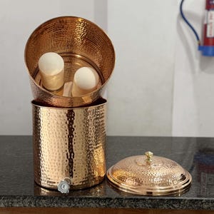 Può includere: Un filtro per l'acqua in rame con una texture martellata. Il filtro comprende una base cilindrica, una sezione superiore con due filtri cilindrici bianchi e un coperchio abbinato. Un rubinetto è visibile sulla base.