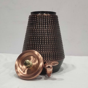 May include: A copper-colored water dispenser with a textured, dotted surface. The dispenser has a matching lid and a spigot. The top and base are dark brown. The lid has a brass knob.