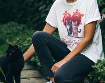 May include: A white t-shirt with a pink heart design featuring two cats. The text "Meow and Forever" is printed below the heart. A person is wearing the shirt and is kneeling on the ground with a black cat sitting in front of them.