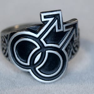Puede incluir: Un anillo de plata con un diseño de esmalte negro de dos círculos entrelazados y una flecha. El anillo es un símbolo de amor y compromiso.
