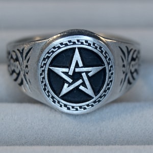Può includere: Un anello d'argento con un designo a pentagramma nero al centro. L'anello ha un bordo decorativo attorno al pentagramma e disegni intricati sui lati.