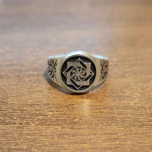 Puede incluir: Anillo de plata con un diseño geométrico de estrella en un marco circular. El anillo tiene patrones decorativos en los lados y un acabado pulido. Una joya.