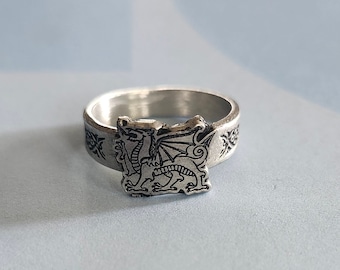 May include: A silver ring with a detailed dragon design. The ring is engraved with a dragon with wings spread out.