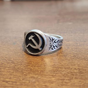 May include: Silver ring featuring a black circle with a white hammer and sickle symbol. The ring has decorative patterns on the sides and is set against a wooden surface. A piece of jewelry with a historical theme.