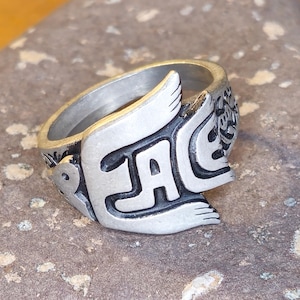 Puede incluir: Anillo plateado con la palabra "PEACE" en letras estilizadas. El anillo presenta diseños en forma de alas y patrones decorativos. El anillo está sobre una piedra gris texturizada.
