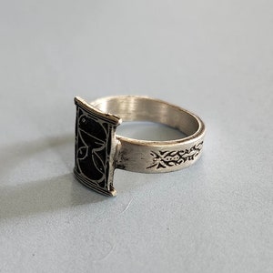May include: A silver ring with a black rectangular design on the front and an intricate design on the band.