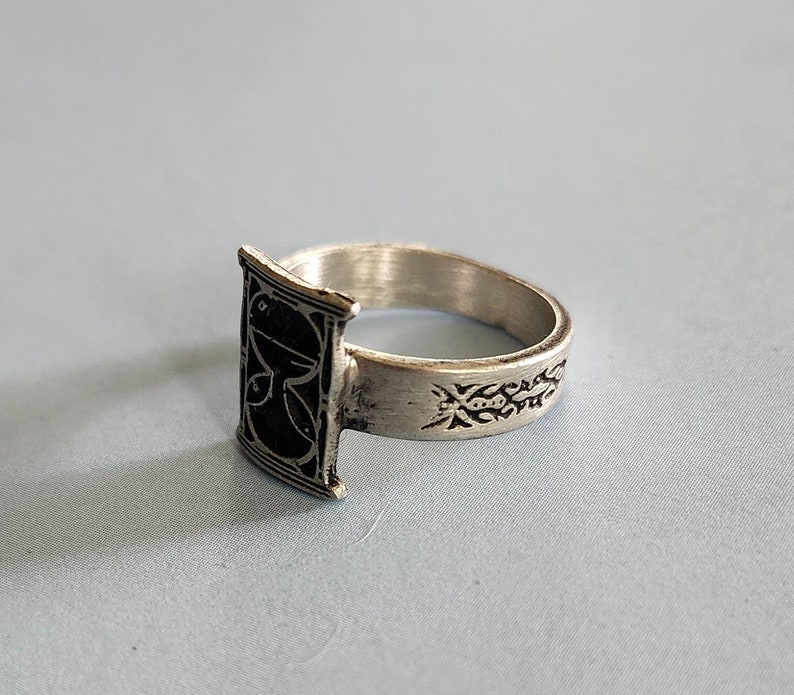 May include: A silver ring with a black rectangular design on the front and an intricate design on the band.
