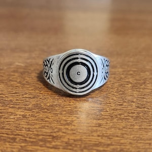 May include: Silver-toned ring with a circular design featuring concentric circles and the letter "C" at the center. The band has decorative patterns on each side. The ring is set against a wooden surface.
