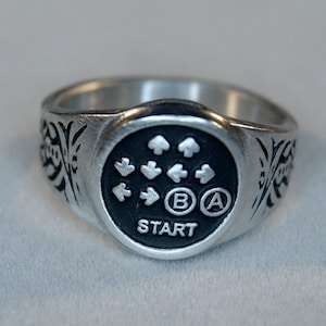 Op de afbeelding: Een zilveren ring met een zwart rond ontwerp met een directionele pad, A- en B-knoppen en het woord "START".