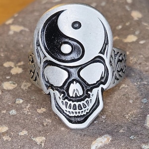 Puede incluir: Anillo plateado con diseño de calavera y símbolo del Yin y Yang en la frente. La calavera tiene rasgos faciales detallados. La banda del anillo tiene un patrón ornamentado. El anillo está sobre un fondo texturizado.