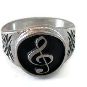May include: A silver ring with a black enamel inlay featuring a treble clef. The ring has a decorative design on the band.