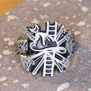 May include: Silver-toned ring with a detailed design featuring a central emblem and ornate side patterns. The ring's design includes a ladder-like structure and other decorative elements. The ring is displayed on a textured, gray surface.