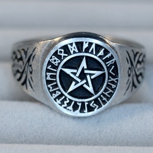 Può includere: Un anello d'argento con un pentagramma nero circondato da rune. L'anello ha un design decorativo sui lati.