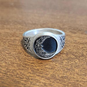 Puede incluir: Anillo plateado con un diseño de media luna sobre un fondo negro, con patrones detallados. El anillo tiene patrones decorativos en los laterales. Una joya con temática celestial.