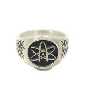 May include: A silver ring featuring a black enamel atom symbol with the letter 'A' inside. The ring has a decorative design on the sides.