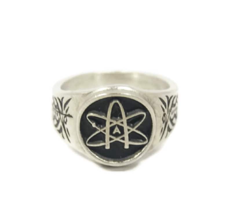 May include: A silver ring featuring a black enamel atom symbol with the letter 'A' inside. The ring has a decorative design on the sides.